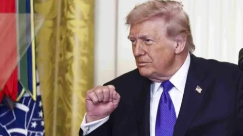 Donald Trump u tamnom odijelu s ljubičastom kravatom ispred američke zastave.