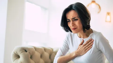 Žena drži ruku na prsima s bolnim izrazom lica u svijetloj prostoriji.