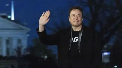 Elon Musk u crnom sakou maše rukom ispred Bijele kuće noću.