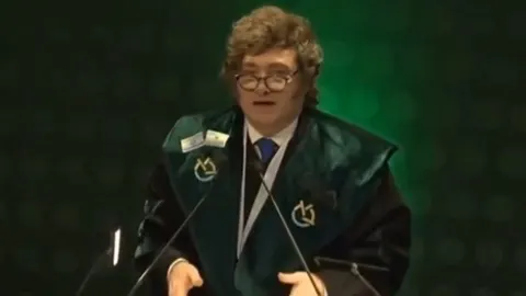 Javier Milei u akademskoj halji s naočalama govori za govornicom.