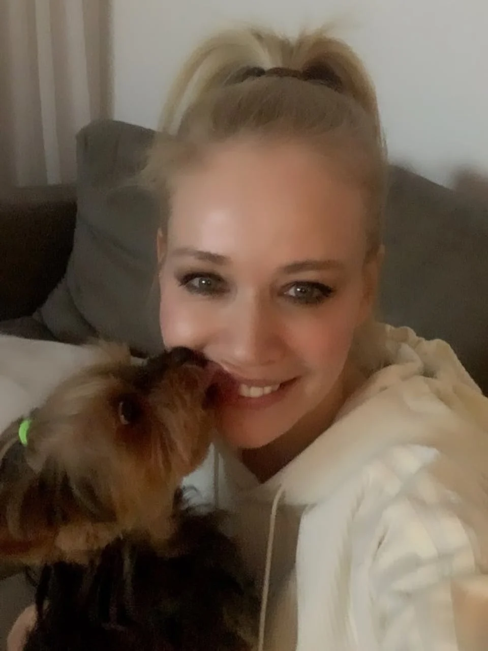 Nasmijana Jelena Rozga u bijeloj majici s kapuljačom dok je njezin pas ljubi u obraz.