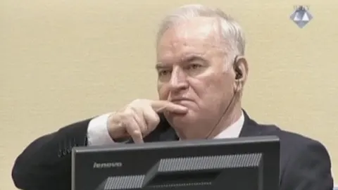 Ratko Mladić u sudnici s prstom na licu i slušalicom u uchu ispred monitora.
