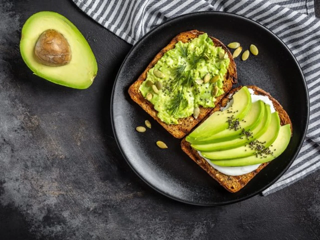 Dva tosta s avokadom i sjemenkama na crnom tanjuru uz cijeli avokado
