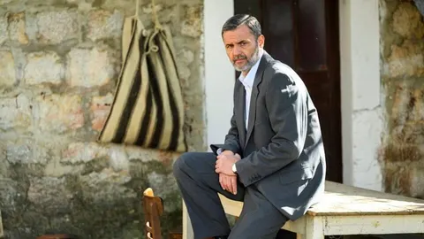 Vladimir Posavec Tušek u sivom odijelu sjedi ispred kamene kuće