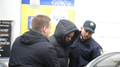 Policajac u odori vodi osobu s kapuljačom preko glave ispred zgrade.