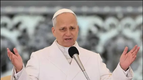 Papa u bijeloj halji drži govor ispred mikrofona s raširenim rukama.