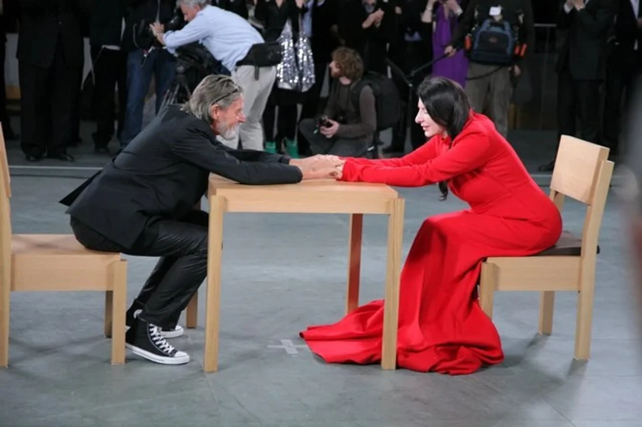 Marina Abramović u crvenoj haljini i Ulay drže se za ruke sjedeći za stolom.