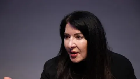 Marina Abramović s tamnom kosom i u crnoj odjeći govori tijekom intervjua.
