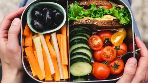 Metalna posuda za ručak s raznim rezanim povrćem i sendvičem od cjelovitih žitarica.