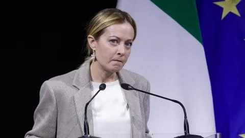 Giorgia Meloni s ozbiljnim izrazom lica stoji za govornicom ispred talijanske zastave.