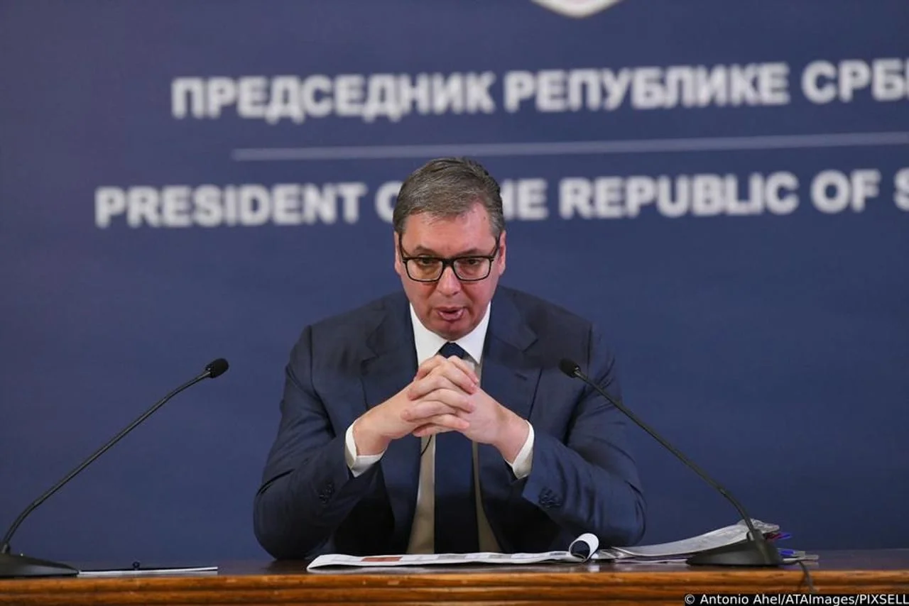 Vučić sjedi za govornicom s mikrofonima ispred plave pozadine s natpisom.