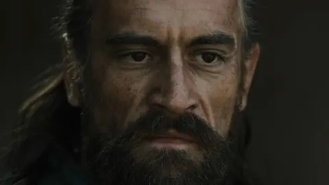 Portret sredovječnog muškarca s gustom bradom i ozbiljnim izrazom lica.