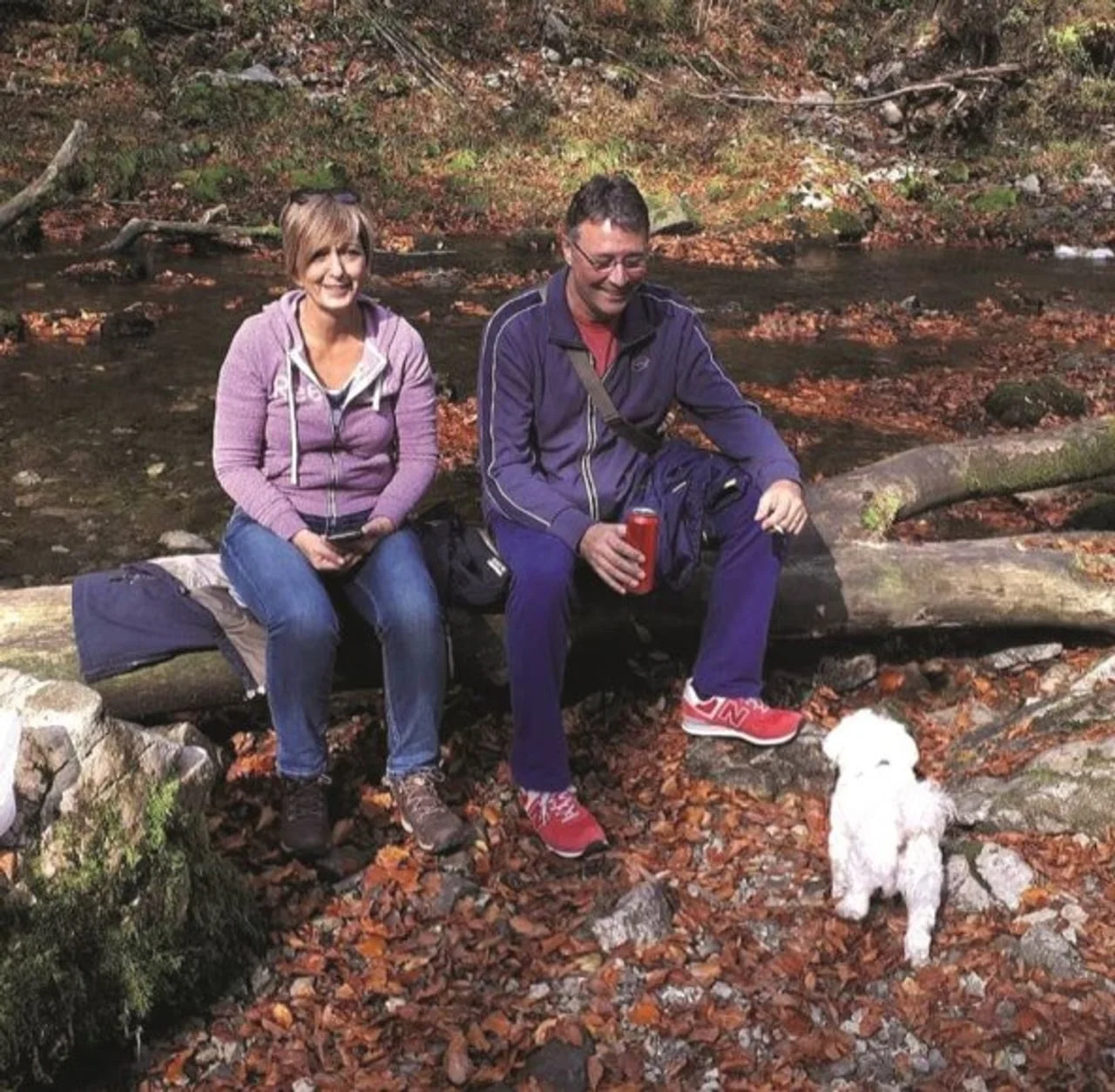 Bračni par Rončević sjedi na srušenom stablu u prirodi pored malog bijelog psa.