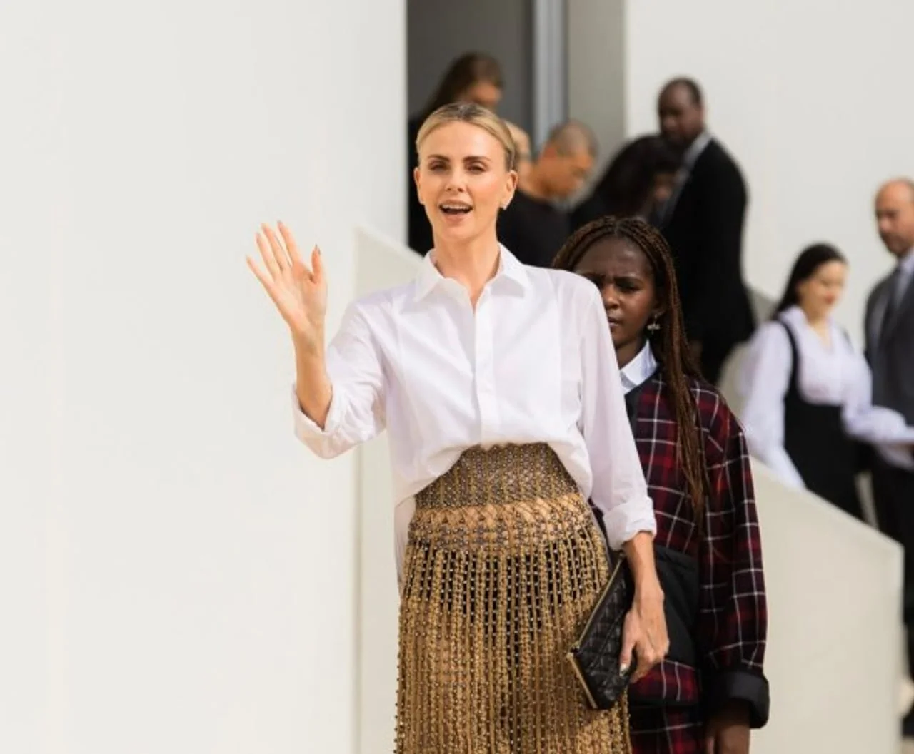 Charlize Theron maše okupljenima noseći bijelu košulju i zlatnu suknju.