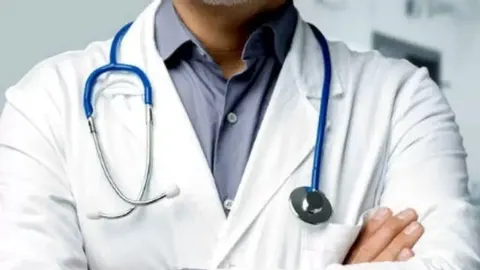 Liječnik u bijeloj kuti s plavim stetoskopom oko vrata i prekriženim rukama.