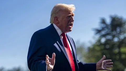 Donald Trump u odijelu s crvenom kravatom gestikulira rukama vani.