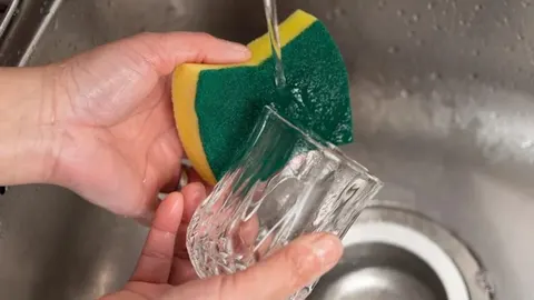 Rukama se pere staklena čaša pomoću žuto-zelene spužve u sudoperu.