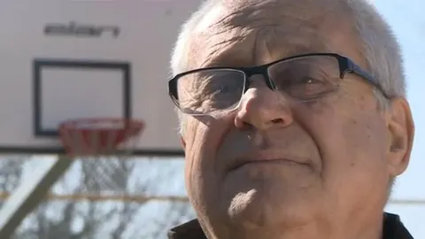 Portret starijeg muškarca s naočalama ispred košarkaškog obruča.