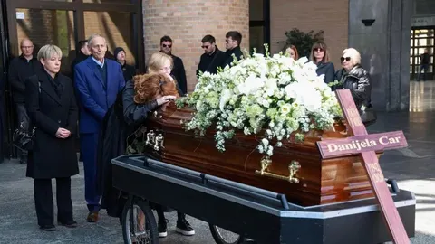 Obitelj i prijatelji pored lijesa prekrivenog bijelim cvijećem na krematoriju.