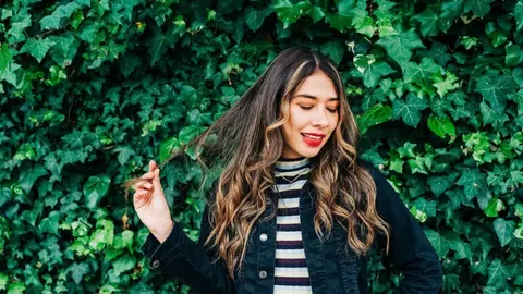 Mlada žena u prugastoj majici i crnoj jakni ispred zida od bršljana.