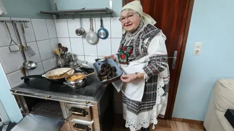 Starija žena u narodnoj nošnji pored peći drži pladanj s domaćim delicijama.