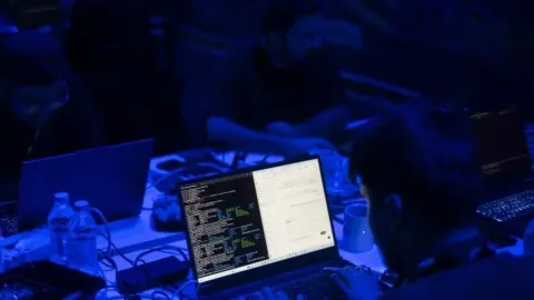 Osoba radi na laptopu s programskim kodom u tamnoj prostoriji s plavim osvjetljenjem