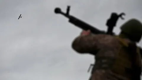 Vojnik u uniformi nišani puškom prema malom dronu na sivom nebu.