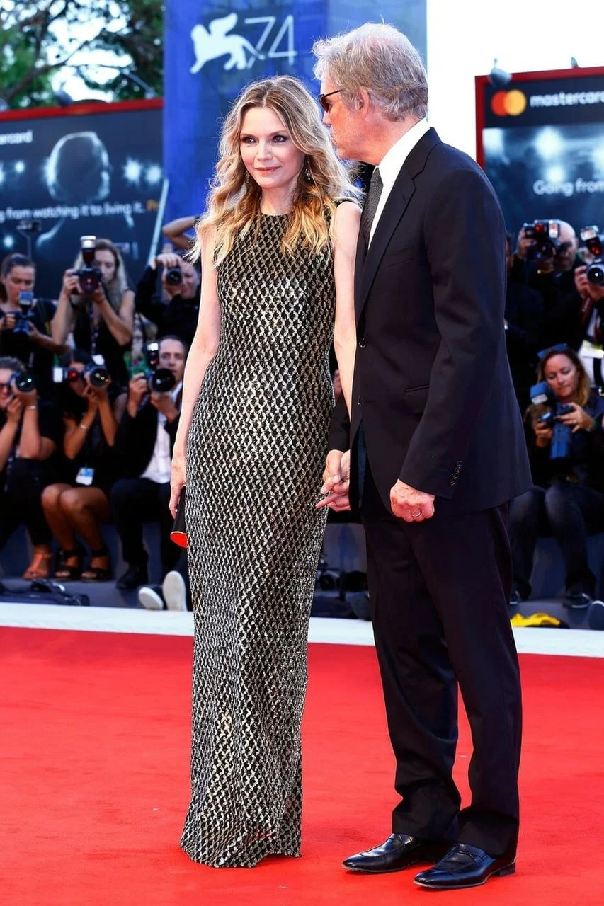 Michelle Pfeiffer u dugoj svjetlucavoj haljini stoji uz supruga na crvenom tepihu.