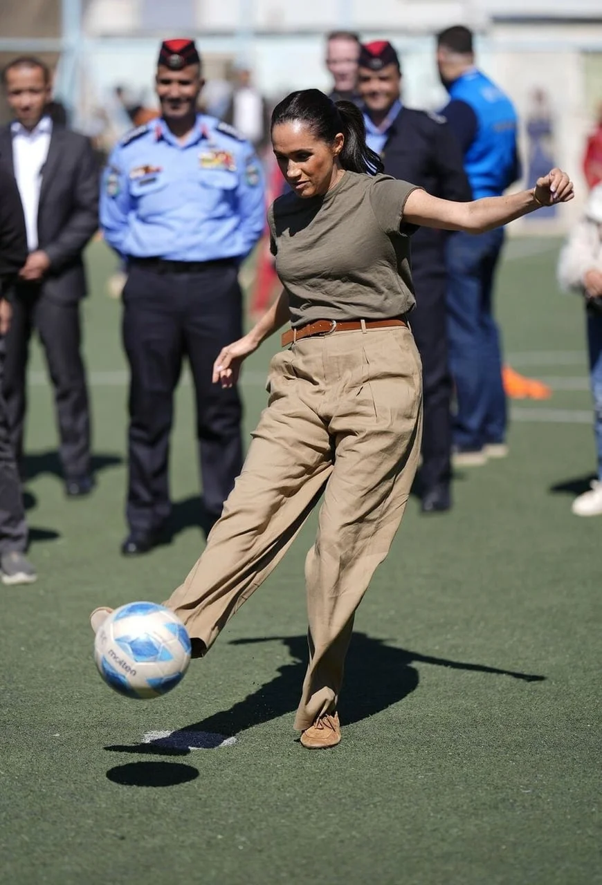 Meghan Markle u bež hlačama udara nogometnu loptu na terenu.