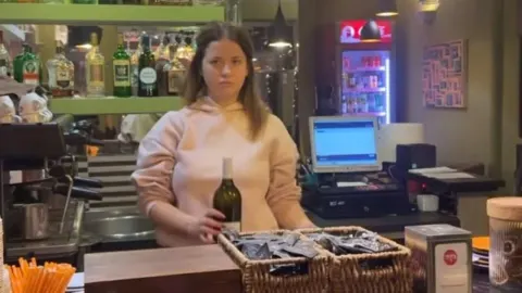 Djevojka u ružičastoj majici drži bocu vina u ugostiteljskom objektu.