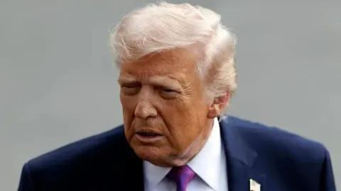 Donald Trump u tamnom odijelu s ozbiljnim izrazom lica na sivoj pozadini.