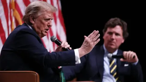 Donald Trump govori u mikrofon dok Tucker Carlson sjedi u pozadini.