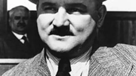 Crno-bijeli portret muškarca s brkovima u tamnom odijelu i sa šeširom.