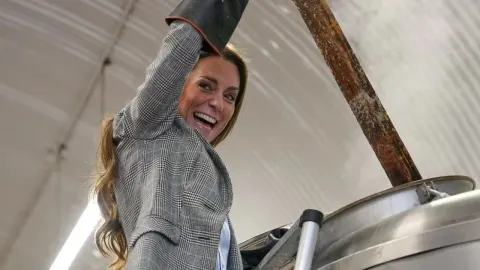 Nasmijana Kate Middleton miješa sadržaj u velikom metalnom kotlu.