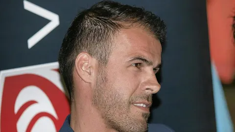 Kratko ošišan muškarac s bradom gleda u stranu ispred tamne pozadine.