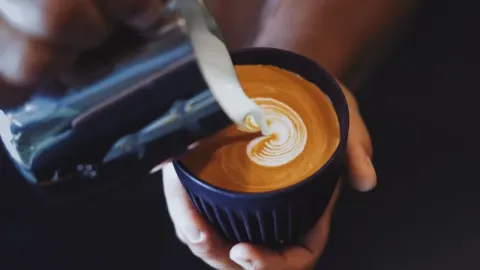 Ruke s vrčem za mlijeko pjene kavu u tamnoj keramičkoj šalici dok se stvara latte art.