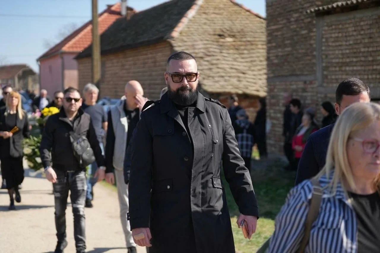 Muškarac s bradom i u tamnom kaputu hoda ispred okupljenih