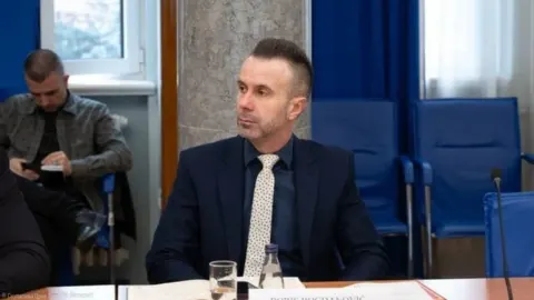 Političar Boris Bogdanović u tamnom odijelu sjedi za govornicom tijekom službenog sastanka.