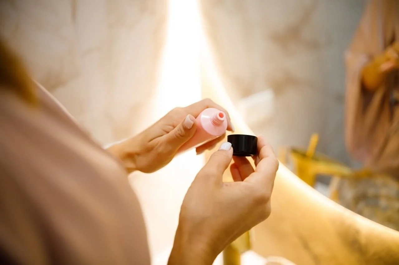 Ruke drže otvorenu ružičastu tubu kreme ili tekućeg pudera iznad umivaonika.