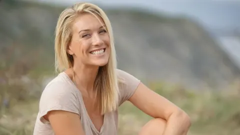 Nasmijana plavokosa žena u četrdesetim godinama sjedi na otvorenom