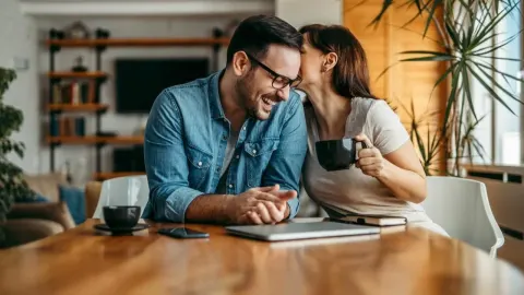 Nasmijani muškarac i žena sjede za stolom dok mu ona šapuće na uho