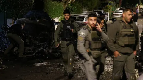 Naoružani vojnici u uniformama pored uništenog crnog automobila noću.