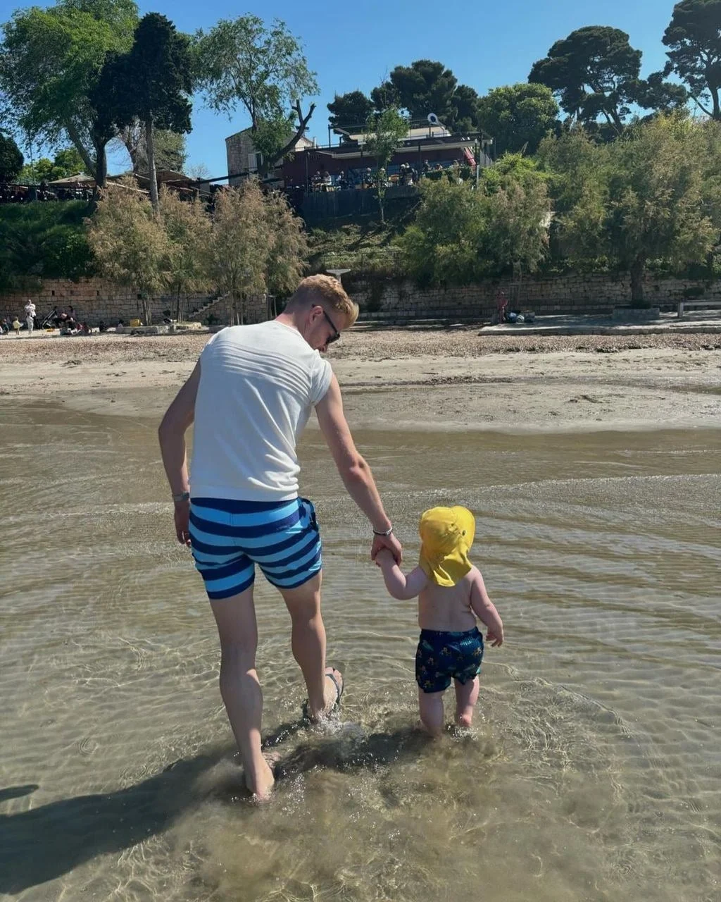 Muškarac i malo dijete hodaju držeći se za ruke u plitkom moru na plaži.