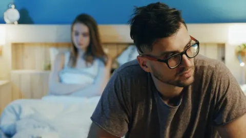 Zabrinuti muškarac s naočalama sjedi na rubu kreveta dok žena u pozadini leži.