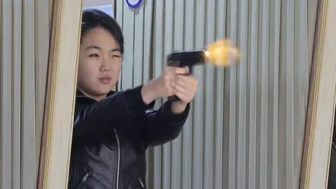 Mlada Kim Ju Ae u kožnoj jakni puca iz pištolja u tvornici oružja.