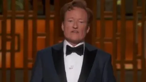 Conan O’Brien u elegantnom smokingu s crnom leptir mašnom na pozornici.